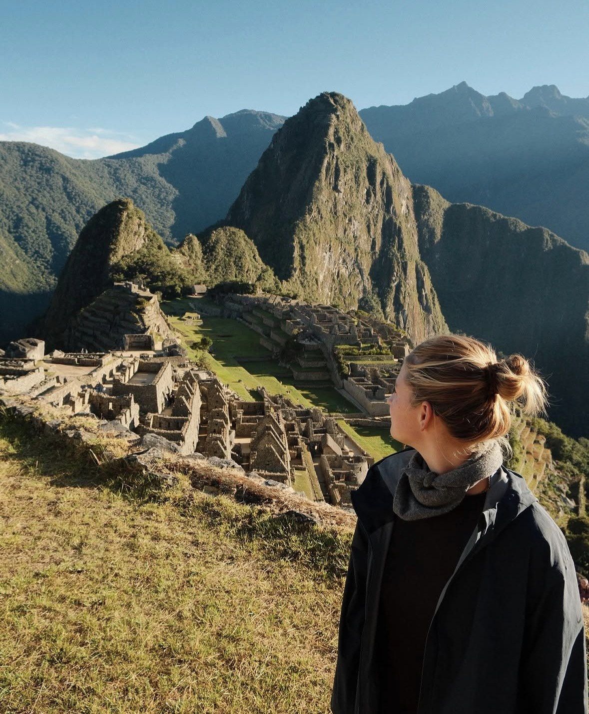 Woman in black jacket watching sunlit Machu Picchu ruins at dawn